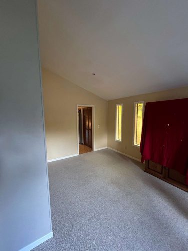A beige carpeted room in Oneal features three tall windows and a doorway. A red cloth covers furniture against one wall. For painting services in Portland, contact Pearl Painters.