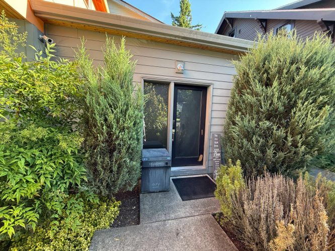 A small gray building in Portland with a glass exterior door and tall shrubs, painted by Pearl Painters and photographed by Arain in May 2025.