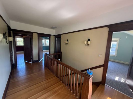 Second-floor hallway with wood floors and trim, wall-mounted lights, and multiple doorways in the Dennis home, Portland. Painted by Pearl Painters as of May 2025.