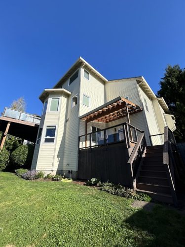 A two-story beige house with multiple windows, a wooden deck with pergola, and backyard stairs at Mackey-Hinzdel, painted by Pearl Painters in June 2025. Located outside Portland.
