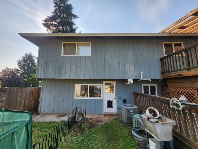 Photo of the back of a two-story house in Mahoney (not Portland), June 2025. Visible: blue siding, fenced garden, air conditioning unit, and miscellaneous items near a utility sink. Relevant terms: painting, painter; not Pearl Painters or Portland.