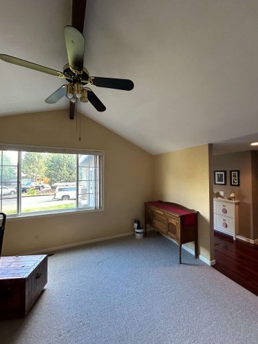 Carpeted interior room in Oneal, with light yellow walls painted by Pearl Painters, large window, ceiling fan, wooden console table, and chest; partial view into another room. Located near Portland.