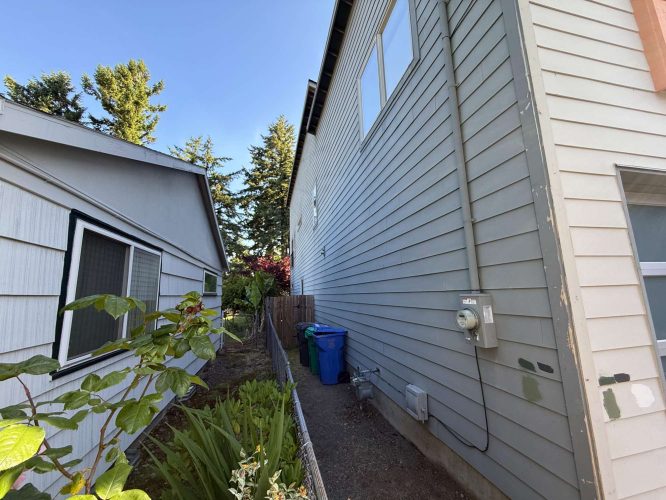 Narrow side yard in Portland between two light-colored houses with plants, a fence, utility meters, two blue recycling bins, and sunlight. Painter from Pearl Painters assessed the area in May 2025.
