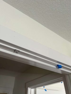A close-up photo shows a painted white interior door frame in Arnold, May 2025. Blue painter’s tape and a patch of spackle are visible above the doorway, likely marking spots for touch-ups by painters or painting contractors.