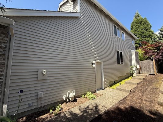 Side view of a Hendricks two-story house in Portland with beige vinyl siding, small garden beds, and a paved walkway. Painting by Pearl Painters. Photo taken May 2025.
