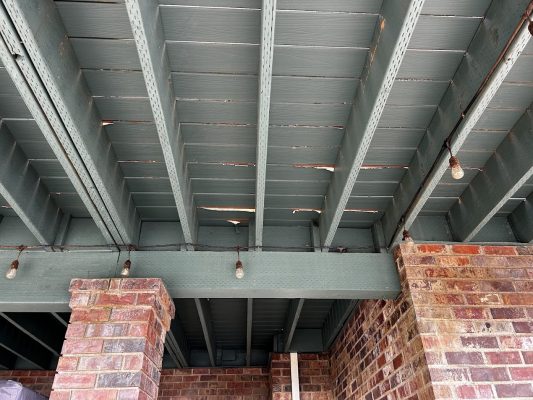 Damaged wooden deck ceiling with cracks, supported by brick columns and string lights, at the Anderson ext in Portland. Suitable for inspection or repair by a painter, such as Pearl Painters. June 2025.