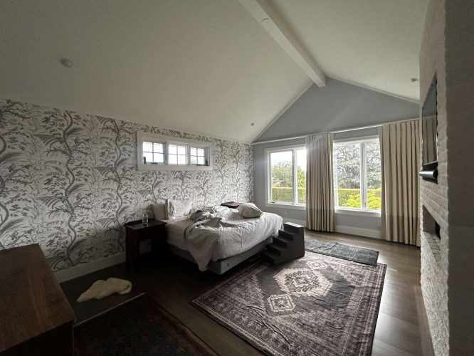 A cozy Portland bedroom painted by Pearl Painters features Duffy floral wallpaper, an unmade bed, large windows with cream curtains, rugs on hardwood floors, and a wooden dresser—ideal for May 2025.