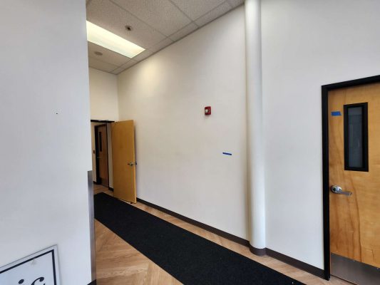 Hallway interior in Portland: white walls painted by Pearl Painters, light wood doors (one open to the Jones office), black mat on floor, fire alarm on wall.