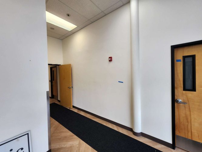 Hallway interior in Portland: white walls painted by Pearl Painters, light wood doors (one open to the Jones office), black mat on floor, fire alarm on wall.