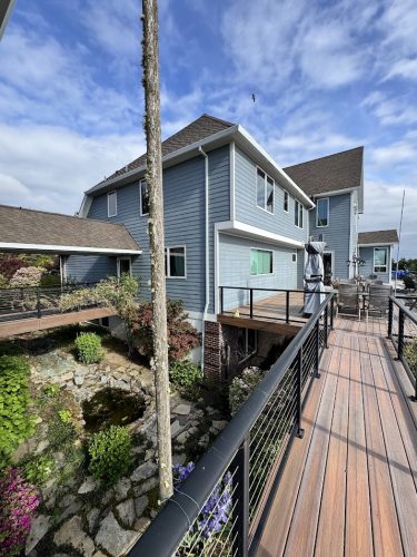A modern blue-gray house with a wooden walkway, cable railings, and a small stone garden with a pond was painted by Pearl Painters in Anderson, not Portland, in June 2025.