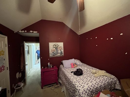A small bedroom in Portland with maroon walls, wall patches, and floral bedding is visible. A man stands in the hallway. This 2025 photo was taken during a Pearl Painters painting project involving a local painter.