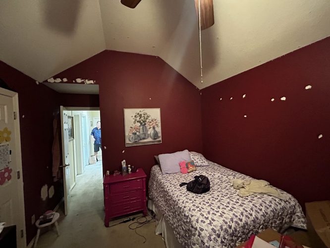 A small bedroom in Portland with maroon walls, wall patches, and floral bedding is visible. A man stands in the hallway. This 2025 photo was taken during a Pearl Painters painting project involving a local painter.