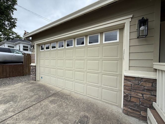 A beige two-car garage door with small windows tops an Emory house with stone and siding. A car and fence are on the left. Photo taken in Portland by Pearl Painters, June 2025.