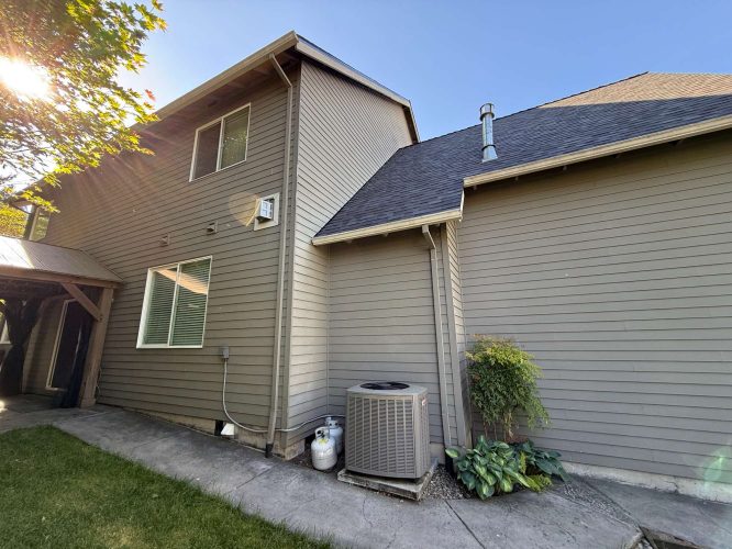 Gray two-story house exterior in Austin shown under a clear May 2025 sky, with visible air conditioning unit, propane tank, and green plants. Example of painting work by Pearl Painters. This project is not located in Portland.