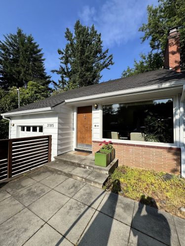 This Carlton single-story house, painted with white siding by Pearl Painters, features a wooden front door, large front window, attached garage, and green planter at the entry. Trees and blue sky frame the June 2025 exterior scene. Location: Carlton (not Portland).