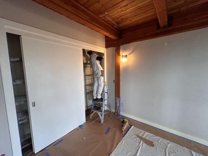Painter in white work clothes on a ladder paints a closet in a room with wooden beams; project by Pearl Painters, Portland.