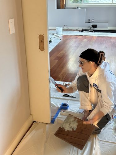 Painter kneeling indoors, painting a wall near a door in Portland; drop cloths cover the floor. Pearl Painters project.