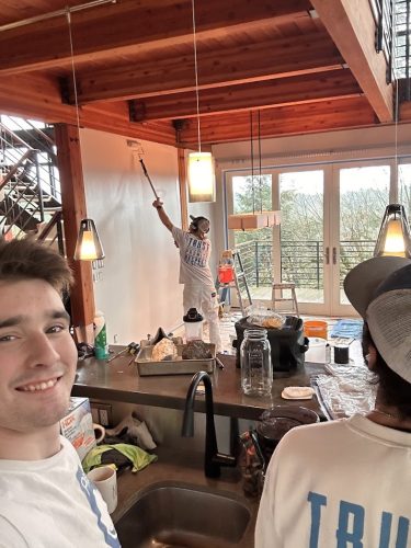 Three painters painting a modern wooden house interior in Portland; supplies on counter, one taking a selfie. Pearl Painters.