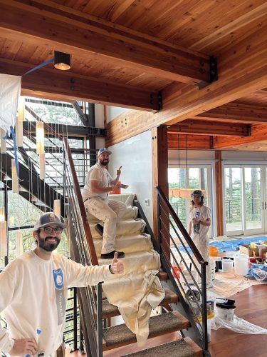 Three painters from Pearl Painters, Portland, posing in a wooden-ceiling interior with painting supplies on stairs and floor.
