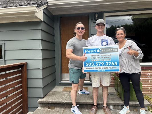 Three people stand outside a Carlton house, holding a Pearl Painters sign for painting services. The sign includes contact details and notes booking into June 2025.