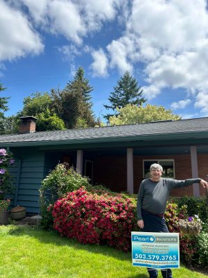 A painter stands in front of a house with blooming bushes in Portland, next to a Pearl Painters sign advertising expert interior and exterior painting services by Persichetti.