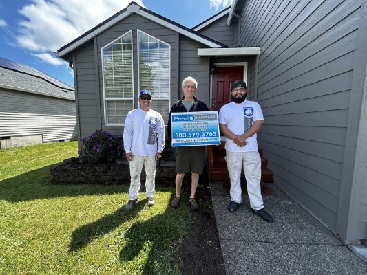 Three painters in uniform stand in front of a gray house in Horn, holding a "Peralt Painters" sign. They are ready for an exterior painting project on a sunny May 2025 day. (Not affiliated with Pearl Painters of Portland.).