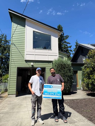 Two painters stand in front of a green house in Portland holding a Pearl Painters sign, displaying their exterior painting completed in May 2025.