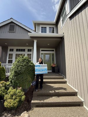 A painter stands on the front steps of a modern Hendricks house in Portland, holding a real estate sign, May 2025. Pearl Painters.