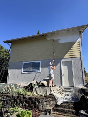 A painter with a roller paints a house exterior white in Portland; tarps protect below. Pearl Painters, July 2025.