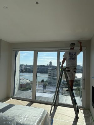 A painter from Pearl Painters paints a ceiling on a ladder in Portland, with large windows showing an industrial waterfront.