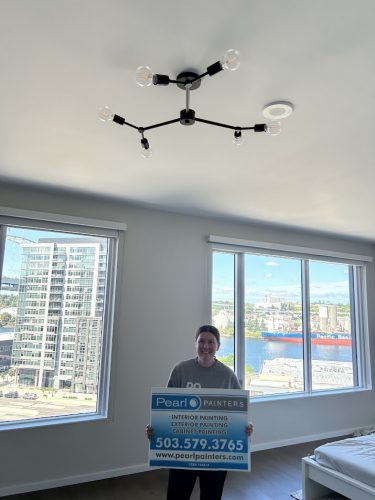 Person holding Pearl Painters sign in a bright interior; Allen cityscape with river and ships visible through large windows.