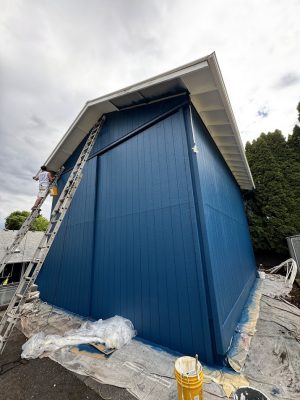 A painter on a ladder paints blue trim; Pearl Painters’ supplies and drop cloths are set out for exterior painting in Portland.