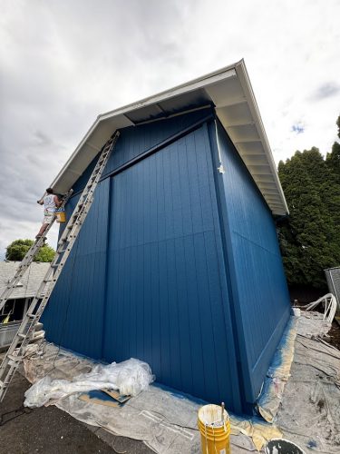 A painter on a ladder paints blue trim; Pearl Painters’ supplies and drop cloths are set out for exterior painting in Portland.