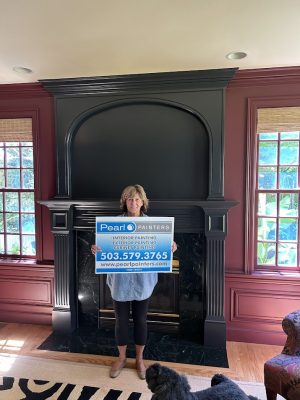 Woman holding Pearl Painters sign for interior and exterior painting services in Purcell stands by black fireplace.