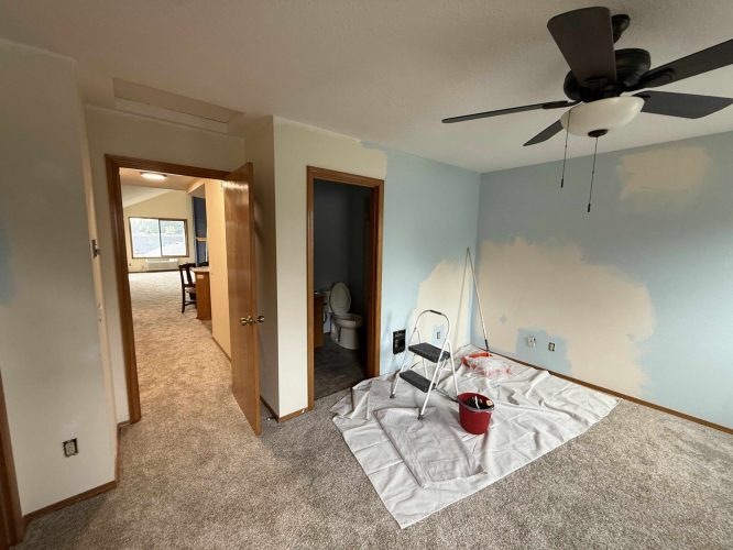 In April 2025, a Yates interior in Portland shows partially painted blue and beige walls. Painting supplies—a step ladder, red paint bucket, and drop cloth—are on the carpet near an open door to a bathroom and hallway. Pearl Painters are handling the project.