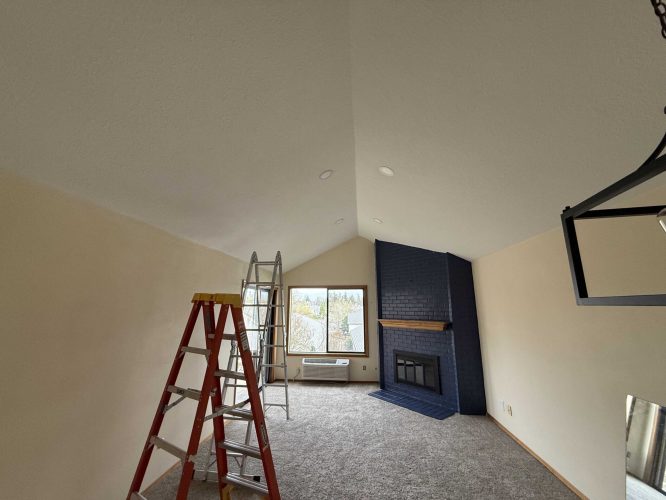 A vaulted ceiling room in Portland features beige walls, a blue fireplace, carpeted floor, and two ladders. Sunlight enters through the windows—ready for repainting by Pearl Painters in April 2025.