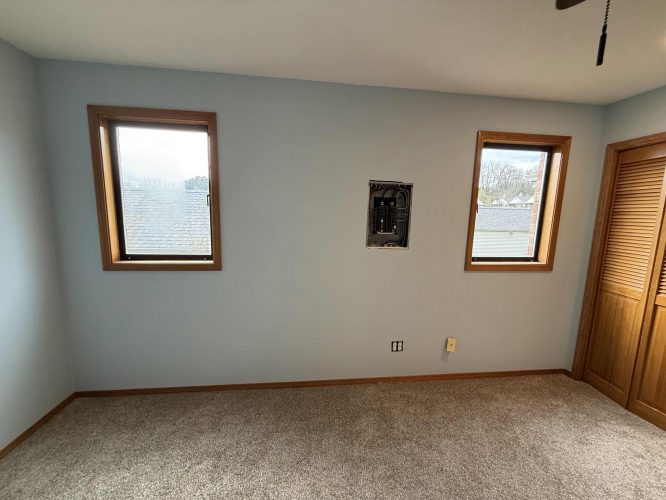 A light blue interior wall with two windows, an exposed electrical panel, beige carpeting, and a wooden closet door at Yates (not in Portland), photographed in April 2025. Painter: Pearl Painters.