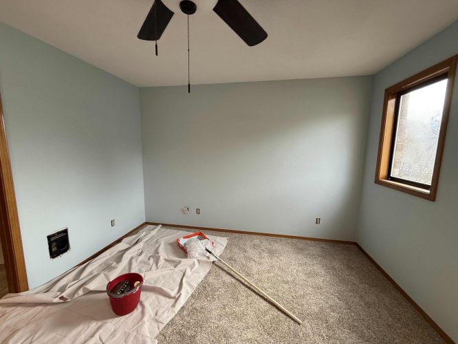 A Yates room in Portland with carpet, light blue walls, a window, and ceiling fan is prepared for painting by Pearl Painters in April 2025, with supplies including a drop cloth, paint roller, and red bucket.