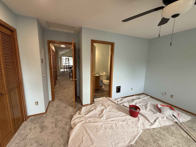 A painter from Pearl Painters is painting a Portland bedroom with light blue walls. Drop cloths, a red bucket, and painting supplies are shown; the adjacent bathroom and hallway are visible. Project completion is scheduled for April 2025.
