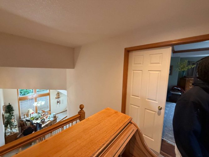 This photo, taken in April 2025, shows a wooden staircase, handrail, closed white door, and part of a living room with large windows and furniture. Mann Interior painting by Pearl Painters in Portland.
