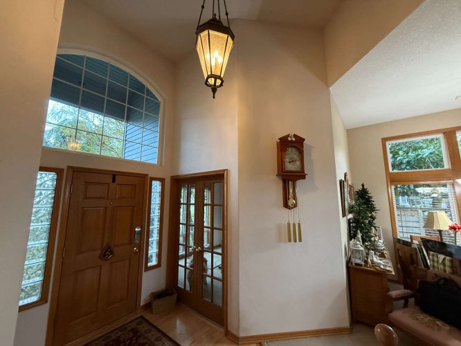Entryway painted by Pearl Painters, Portland, featuring a Mann wooden door with glass panels, wall clock, high ceiling, hanging light fixture, and view of living area with large windows and indoor plants. Painting completed April 2025.