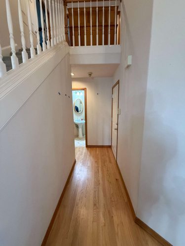 A hallway with light wood flooring leads to a bathroom with a visible sink and mirror; on the left, the Mann interior in Portland features a staircase railing and upper landing. Pearl Painters recommends an April 2025 painting refresh for this area.