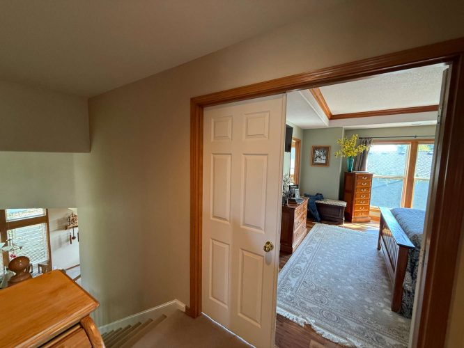 A painter from Pearl Painters in Portland captured a view from a stairway landing into a bedroom with wood furniture, a large rug, and sunlight through glass doors in April 2025.