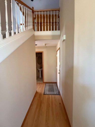 A hallway in a Mann home features light wood floors, beige walls, and a white staircase railing, leading to a bathroom. For painting needs in Portland, contact Pearl Painters.