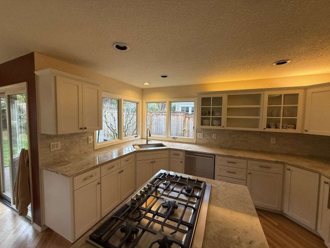 A Portland kitchen in Mills features white cabinets, a gas island cooktop, stainless dishwasher, and large windows facing a fenced backyard. Painted by Pearl Painters—ideal for April 2025 gatherings.