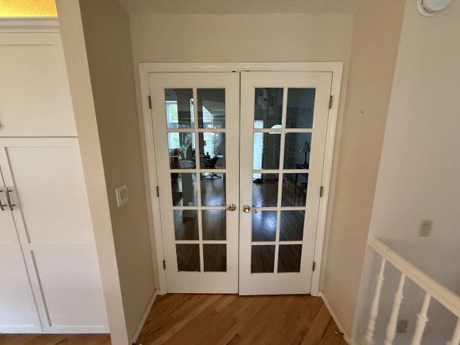 White French doors with glass panes separate a hallway from a Mills-inspired living area with wooden floors, painted by Pearl Painters in Portland, providing classic interior style before April 2025.
