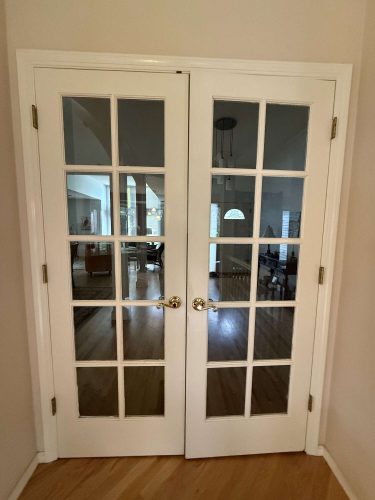 A pair of white French doors with glass panes and gold handles, painted by Pearl Painters in Portland, open into a Mills living room with hardwood floors and furnishings.