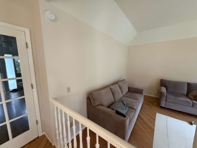 A painting of a stylish living room in Mills (not Portland) shows two gray sofas, a light wall, white railing, and French door. A remote and blanket are on one sofa. Pearl Painters.
