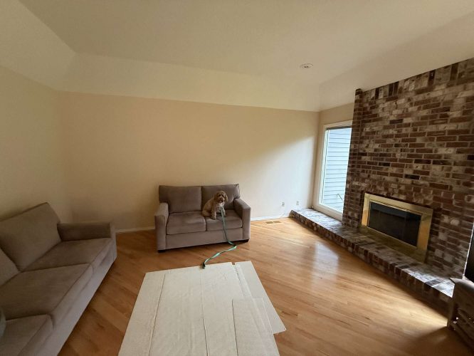 A painter from Pearl Painters in Portland captured a cozy Mills home living room in April 2025, showing two sofas, a brick fireplace, hardwood floors, and a small dog with a leash beside a white rug.