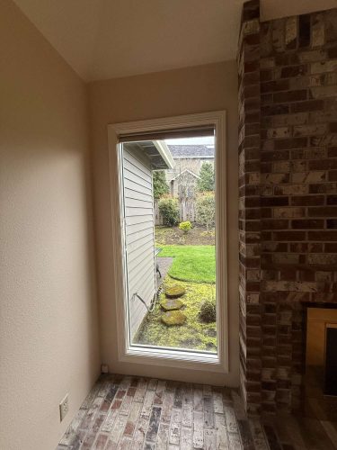This painting by a Portland painter from Pearl Painters shows a garden in Mills in April 2025, viewed through a large window beside a brick fireplace.
